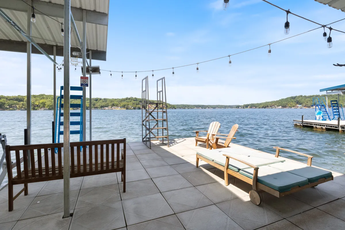 A waterfront lounging area with sunbathing chairs and a wooden bench sits at the water's edge, accented by string lights for evening ambiance. The clear lake invites swimming, and a ladder provides access for water activities. The tranquil setting is perfect for sunbathing, fishing, or enjoying playful afternoons by the lake.