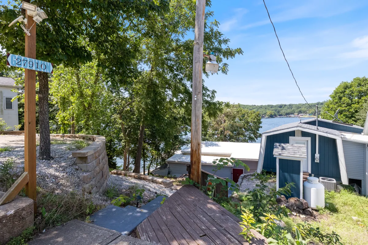 A tranquil scene featuring a blue house with a wooden pathway leading to the water, flanked by a signpost and surrounded by greenery. The view emphasizes easy access to nature and water activities.