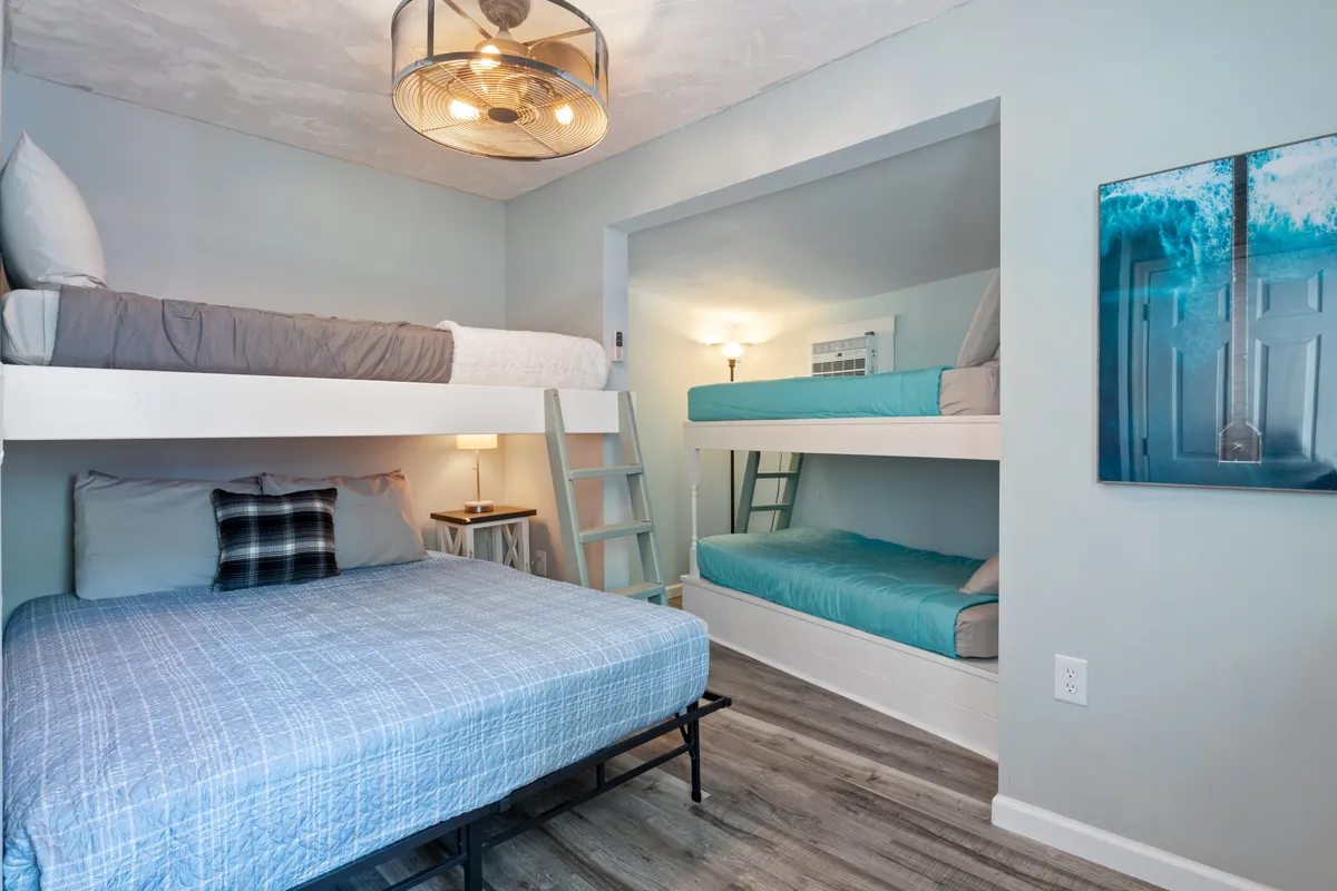 A family-friendly bedroom with both bunk beds and a queen bed, highlighted by a decorative light fixture and plaid accents. Large windows allow for ample natural light, creating a warm and relaxed atmosphere ideal for families or groups of friends.