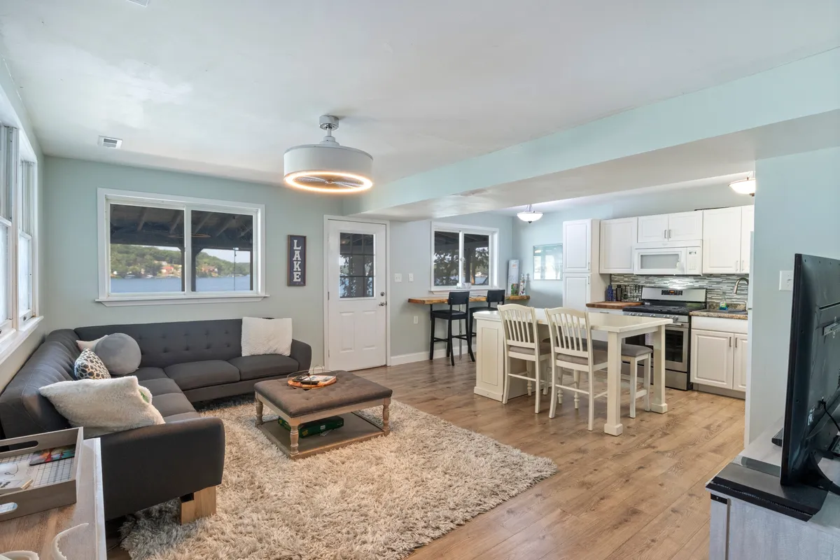 An open-concept living and dining area features a plush sofa, a coffee table, and a dining table set near the kitchen. Warm, golden light streams through the windows, hinting at sunset views. The layout fosters seamless transitions between socializing, dining, and relaxing, making it ideal for hosting or unwinding after lakeside adventures.