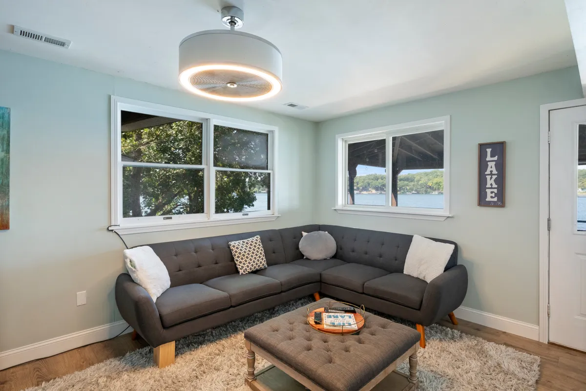 A cozy sectional sofa faces expansive windows that overlook the lake, allowing natural light to flood the living area. The space is styled with a modern light fixture and a decorative coffee table, creating an inviting environment for relaxation or conversation. The tranquil water view is a dominant feature, emphasizing the connection between indoor comfort and the scenic outdoors.