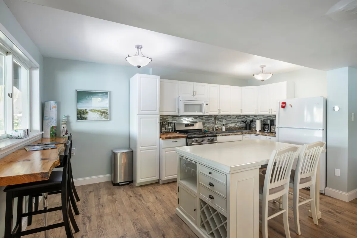 This image features a bright kitchen space with a central kitchen island, white cabinetry, and stainless steel appliances. Bar stools line the island, making it ideal for both casual breakfasts and group meal prep. The overall atmosphere is welcoming and designed for gathering.
