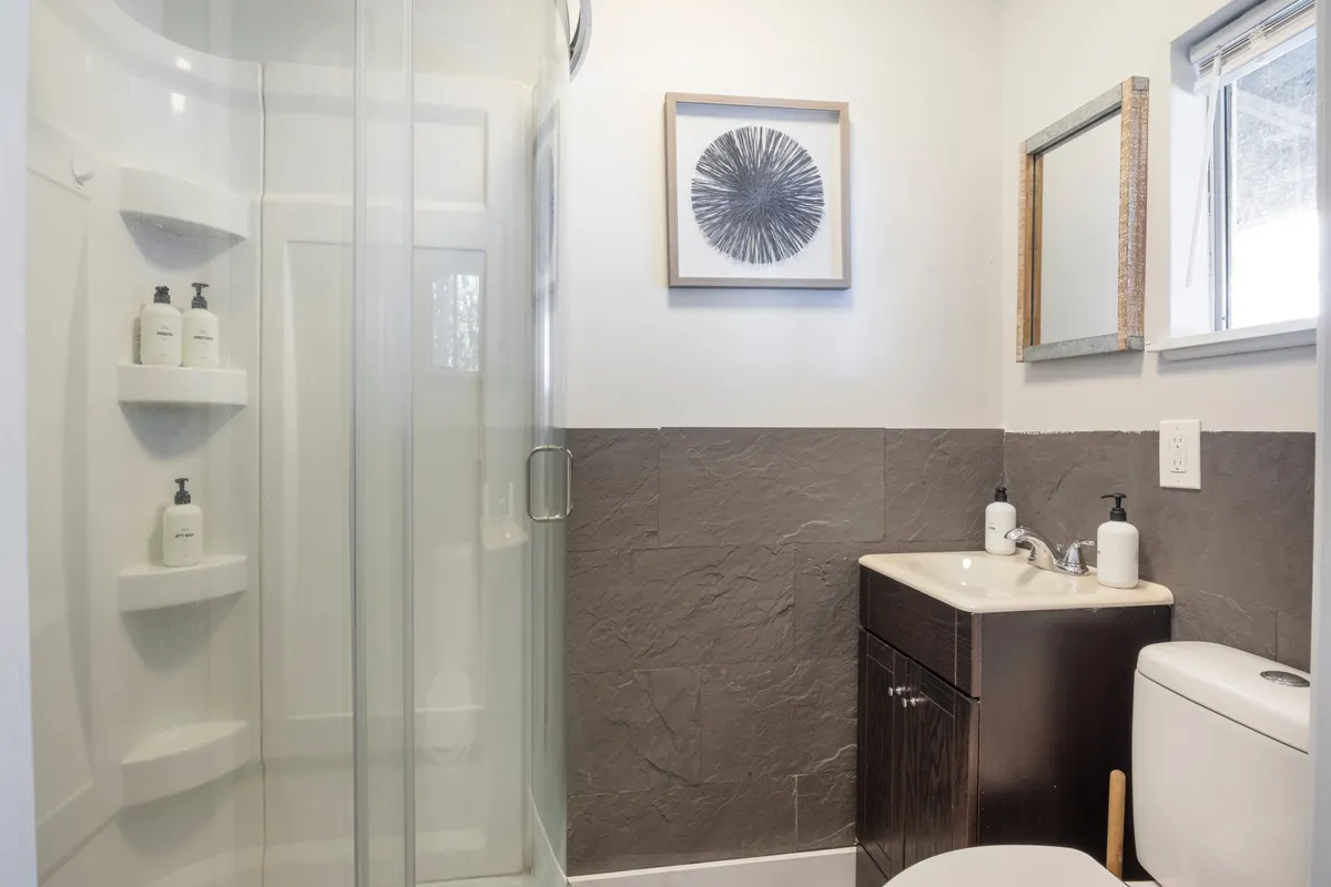 A modern bathroom featuring a glass shower enclosure, sleek sink and vanity, and artwork. Natural light streams in through a window, emphasizing the space's calm and fresh atmosphere—perfect for relaxing after a day by the lake.