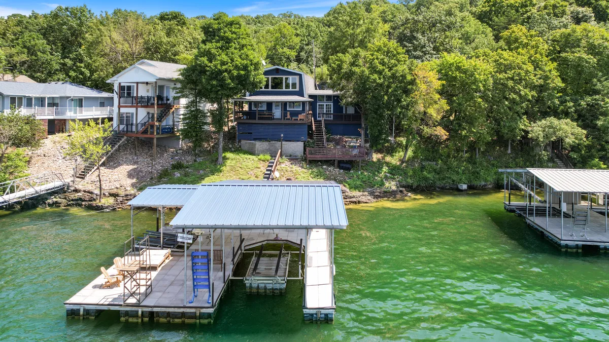 A tranquil shot of the lakefront dock extending out over calm waters, with the main house nestled among surrounding trees in the background. The serene setting emphasizes private lake access and a peaceful atmosphere ideal for families.