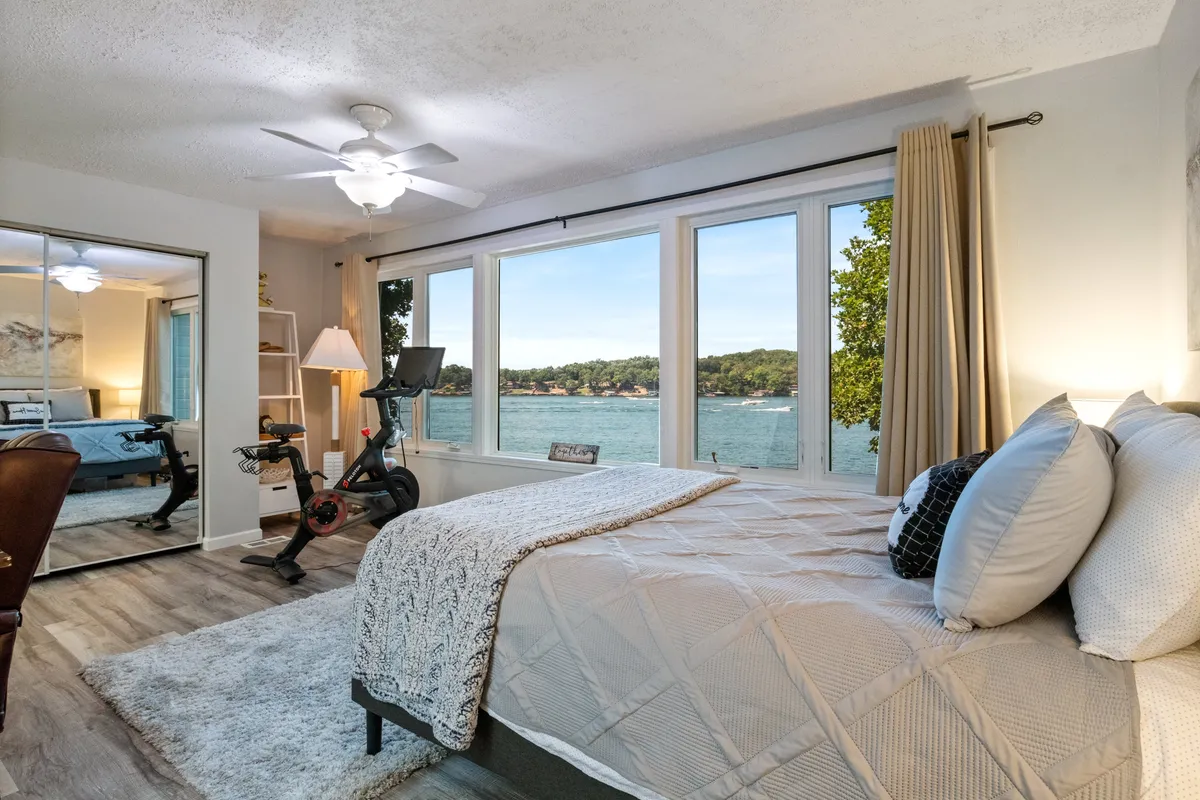 A tranquil bedroom featuring large windows that frame a beautiful lake view. The bed is adorned with decorative pillows, and an exercise bike and wooden chair are positioned to take advantage of the scenery, offering both relaxation and activity in a peaceful setting.