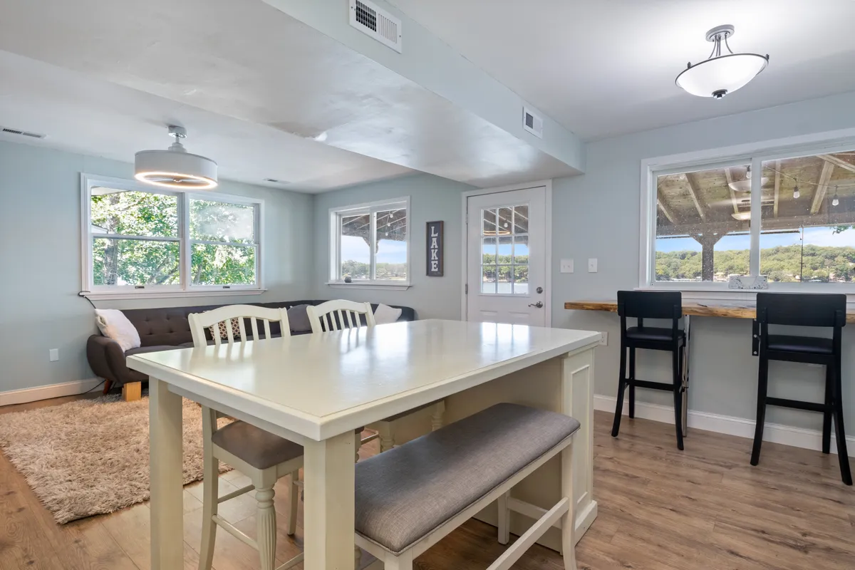 A bright, welcoming space features a dining table adjacent to a comfortable sofa, with large windows offering expansive views of the lake. A bar seating area completes the scene, underscoring the space’s suitability for both family meals and casual entertaining with friends, all while basking in natural light and beautiful scenery.